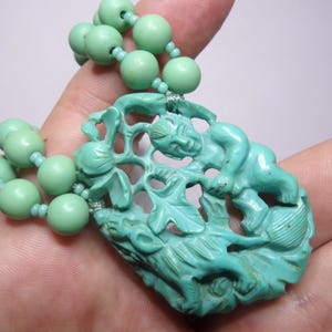 May include: A turquoise green necklace with a carved pendant featuring a mythical creature and a floral design.