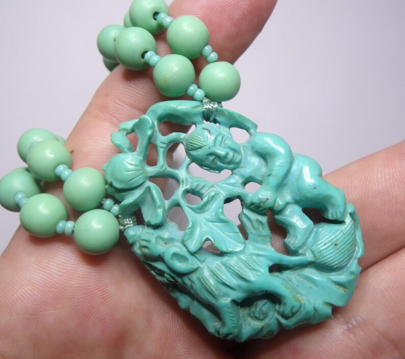 May include: A turquoise green necklace with a carved pendant featuring a mythical creature and a floral design.