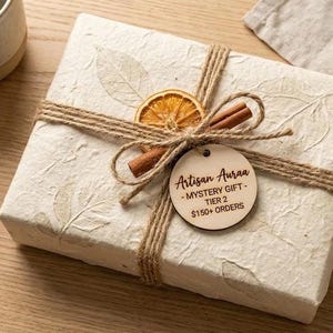May include: A wrapped gift box with a natural, textured paper. It is tied with twine and adorned with a dried orange slice, cinnamon stick, and a wooden tag that reads "Artisan Auraa - MYSTERY GIFT - TIER 2". A ceramic mug sits nearby.