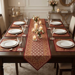 May include: A formal dining table set for a meal. The table is covered with a red and gold patterned runner and placemats. White plates, gold silverware, and clear wine glasses are set for each place. Candles and floral arrangements add to the ambiance.