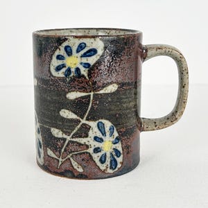 May include: A ceramic mug with a cylindrical shape and a handle. The mug features a dark brown and black background with a floral design in blue, white, and yellow. The mug is suitable for hot or cold beverages.