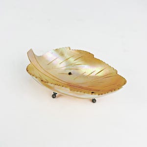 May include: A decorative leaf-shaped dish made of iridescent mother-of-pearl. The dish has a scalloped edge and is supported by three small, dark metal feet. The leaf design features etched veins and a natural, pearlescent sheen.