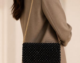 Op de afbeelding: Zwarte kralen clutch met een gouden ketting. De rechthoekige tas is bedekt met kleine, glanzende zwarte kralen. De gouden ketting is dun en delicaat. De tas wordt over de schouder gedragen.