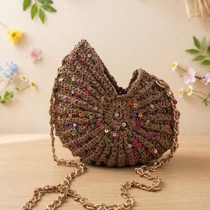 May include: A brown crocheted handbag with colorful sequins. The bag has a unique shape and a gold chain strap. The bag is sitting on a wooden surface. The bag is a stylish accessory.