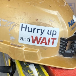 May include: A gold-colored hard hat with a sticker that reads "Hurry up and WAIT" in black and red text. The hard hat has some wear and tear, suggesting it's been used on a construction site.