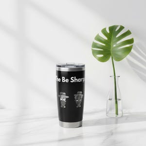 May include: A black stainless steel tumbler with the text "The Be Sharp" in white. The tumbler features two cartoon characters wearing hats and glasses. A green plant in a clear vase is in the background.