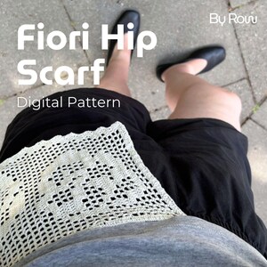 Könnte beinhalten: Ein cremefarbenes gehäkeltes Hüfttuch mit quadratischem Blumenmuster, getragen über einer schwarzen Shorts und einem grauen Shirt. Der Text "Fiori Hip Scarf Digital Pattern" und "By Row" ist eingeblendet, dazu schwarze flache Schuhe.