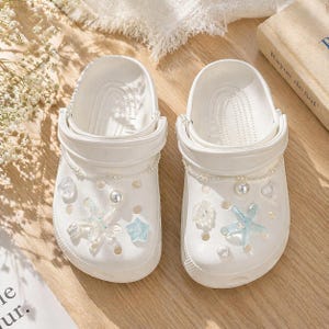 May include: A pair of white clogs decorated with clear and light blue starfish, shells, and pearl accents. The clogs have a strap and are adorned with a pearl chain. The shoes are on a wooden surface.