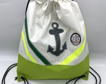 May include: White drawstring backpack with a green bottom and accents. Features a dark gray anchor graphic and a circular patch with text. Includes black drawstrings and green and yellow stripes.