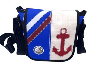 Può includere: Una borsa a tracolla blu e bianca con una grande applicazione a forma di ancora rossa sulla patta anteriore. La borsa presenta un dettaglio a righe diagonali bianche e bordeaux, una tracolla nera regolabile e una toppa circolare con il testo KEL.
