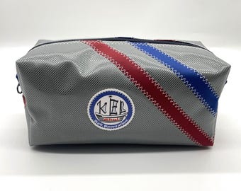 May include: Gray rectangular bag with a zipper closure, featuring red and blue diagonal stripes. A circular patch with the text "KEL Sail Generation" is on the front. The bag is made of a textured material.