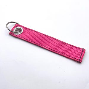 May include: A bright pink fabric keychain strap with black stitching and a silver metal ring. The strap is rectangular and has a reinforced eyelet for the ring. The background is white.