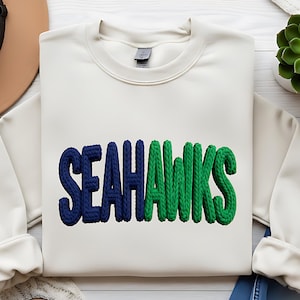 May include: Cream-colored sweatshirt with the word "SEAHAWKS" in navy blue and green, featuring a textured, rope-like design. The sweatshirt is displayed with a brown hat, sandals, and denim shorts.