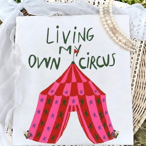 Puede incluir: Lienzo blanco con el texto "Living My Own Circus" en verde. Una carpa de circo rosa y roja con detalles de estrellas está centrada debajo del texto. Una bandera roja está encima de la carpa. El lienzo está decorado con un collar de perlas y encaje blanco.