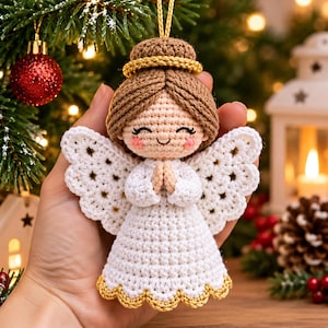 Könnte beinhalten: Ein gehäkelter Engel-Anhänger mit hellbeigem Gesicht, rosa Wangen, braunem Haar in einem Dutt und einem goldenen Heiligenschein. Die Figur trägt ein weißes Kleid mit goldenem Wellensaum und hat weiße gehäkelte Flügel sowie gefaltete Hände.