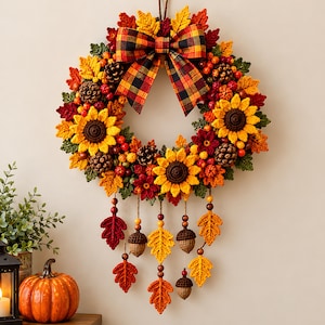 May include: A decorative autumn wreath featuring crocheted sunflowers, maple leaves, pinecones, and red berries. A large plaid bow sits at the top. Five hanging strands with crocheted leaves, acorns, and wooden beads dangle from the bottom.
