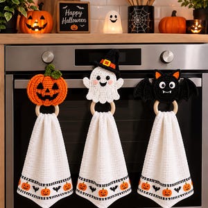 Op de afbeelding: Drie witte wafelstructuur keukendoeken hangen aan een ovenhandgreep, elk voorzien van een houten ring en een gehaakt Halloween-figuur: een oranje pompoen, een wit spookje met een heksenhoed en een zwarte vleermuis met uitgespreide vleugels.