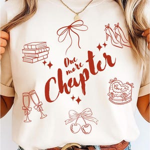 Pode incluir: Camiseta creme com um design gráfico vermelho. O design inclui o texto "One more Chapter" com ilustrações de livros, saltos altos, taças de champanhe, cerejas, um bolo e laços. A camiseta é usada com jeans azuis e um cinto marrom.