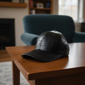 Może przedstawiać: Czarna czapka bejsbolowa z teksturowanym wzorem przypominającym skórę krokodyla. Czapka wykonana jest z ciemnego materiału i leży na drewnianym stole. Czapka ma zakrzywiony daszek i strukturalną koronę.