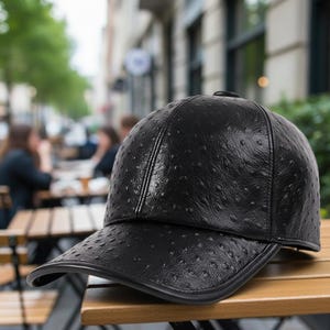May include: Black baseball cap with a textured, dotted pattern. The cap features a curved peak and a button on top. The hat is made of a leather-like material and rests on a wooden surface.