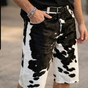 Pode incluir: Calções de ganga com estampado de pele de vaca, cinto de couro preto e fivela prateada. Os calções apresentam um padrão de pele de vaca a preto e branco. Os calções têm uma bainha crua.