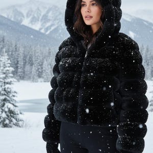 Puede incluir: Chaqueta negra con capucha de piel sintética con cierre de cremallera. La chaqueta tiene un diseño acolchado y se usa con leggings y guantes negros. El fondo presenta un paisaje montañoso nevado.