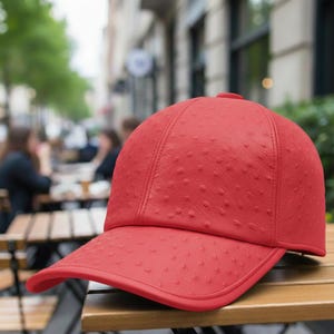 以下が含まれることがあります： 赤色の野球帽。表面はテクスチャ加工が施されています。つばはカーブしており、クラウンは丸みを帯びています。木製のテーブルの上に置かれています。