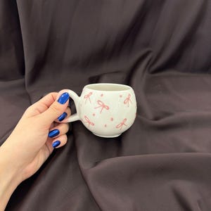Puede incluir: Una taza de cerámica blanca con forma redondeada y asa. La taza está decorada con un patrón de pequeños lazos rosas. La taza está sostenida por una mano con esmalte de uñas azul.