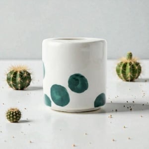 Op de afbeelding: Een witte cilindervormige keramische plantenbak met tealkleurige stippen. Kleine groene cactussen met stekelige naalden omringen de plantenbak. Het ontwerp is minimalistisch en modern, geschikt voor interieurdecoratie.