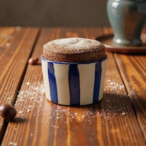 Op de afbeelding: Een chocoladesoufflé, bestrooid met poedersuiker, in een keramische kom met blauw-witte strepen. Het dessert staat op een houten tafel met strooisuiker en een paar hazelnoten. Op de achtergrond staat een blauwe keramische vaas.