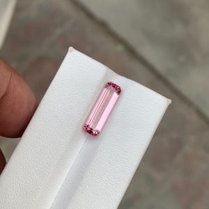 Könnte beinhalten: Ein rechteckiger Edelstein im Baguette-Schliff mit hellrosa Farbton und facettierten Oberflächen. Der Stein zeigt eine klare, durchscheinende Qualität mit tieferer rosa Sättigung an den oberen und unteren Kanten, platziert auf einem weißen, strukturierten Ständer.