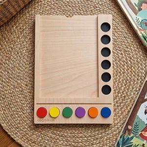 Peut inclure: Jeu éducatif en bois avec un cadre rectangulaire et sept cercles noirs. La base comporte six cercles colorés : rouge, jaune, vert, violet, orange et bleu. Le mot "cognikids" est imprimé sur la base. Un panier tressé et des livres sont en arrière-plan.