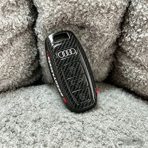 Puede incluir: Llavero de coche de fibra de carbono con el logotipo de Audi y el texto "SPEEDTRACKS" en blanco. El llavero tiene botones para bloquear, desbloquear y abrir el maletero. Se ven detalles en rojo.