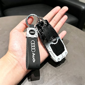 Pode incluir: Porta-chaves de carro a preto e branco com o logótipo da Audi. O porta-chaves está preso a uma correia preta com o logótipo e o texto da Audi. O porta-chaves é prateado.