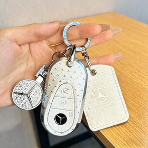 May include: A white car key fob and key ring set with a Mercedes-Benz logo. The key fob is white with a pattern of small silver stars. A round charm and a tag with the Mercedes-Benz logo are also included.