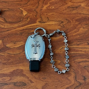 Könnte beinhalten: Ein silberner Autoschlüssel mit Kreuzmuster und ein passendes Kettenarmband. Der Schlüssel hat einen schwarzen Sockel und ein dekoratives Kreuzemblem. Das Armband verfügt über kleine, verzierte Charms und Glieder. Die Artikel werden auf einer Holzoberfläche präsentiert.