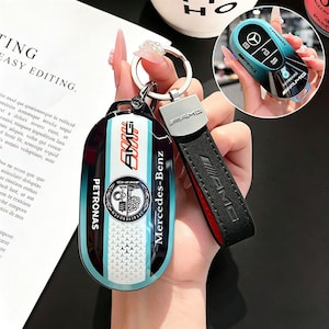 Puede incluir: Una funda para llavero Mercedes-Benz AMG negra, verde azulado y blanca con un llavero a juego. La funda presenta el logotipo de AMG, la marca Petronas y una correa de cuero con el logotipo de AMG.