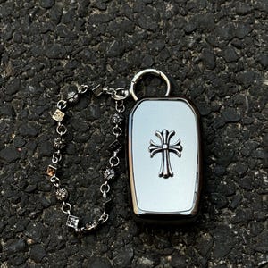May include: A silver key fob case with a cross design, attached to a decorative chain with floral and geometric charms. The case has a polished finish and a metal ring for attachment.