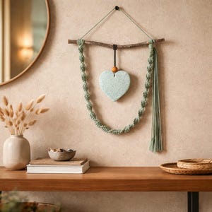 May include: A wall hanging featuring a wooden branch, macrame cord, and a heart-shaped ornament. The macrame is a light green color, and the heart is a pale blue-green. The wall hanging is displayed on a beige wall.