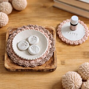 May include: A wooden tray holds a white ceramic plate with three small, round, white objects with leaf designs. The plate sits on a crocheted doily. A small essential oil bottle sits on a matching coaster with the word "PAUSE".