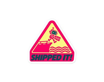 May include: A triangular sticker with a yellow background and a pink border. It features a small robot jumping off a dock into wavy water, holding a box with a code symbol. The text SHIPPED IT! is printed in bold dark blue letters at the bottom.