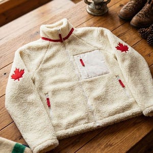 Può includere: Giacca in pile color crema con dettagli rossi e collo alto. Presenta una foglia d'acero rossa sulla manica e una tasca bianca. La giacca ha una cerniera completa e tiralampo rossi. Un capo di abbigliamento caldo.