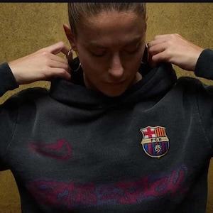 Pu&ograve; includere: Felpa con cappuccio grigio scuro con stemma del Barcellona sul petto. Lo stemma presenta uno scudo con croce, strisce e un pallone da calcio. La felpa ha un aspetto vintage sbiadito.