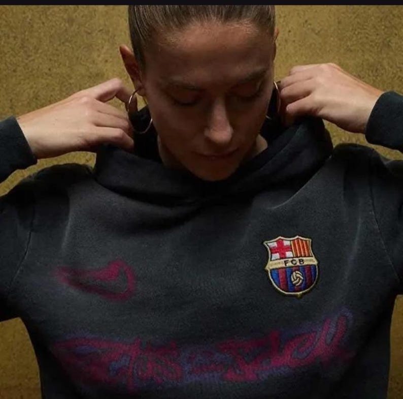 Pu&ograve; includere: Felpa con cappuccio grigio scuro con stemma del Barcellona sul petto. Lo stemma presenta uno scudo con croce, strisce e un pallone da calcio. La felpa ha un aspetto vintage sbiadito.