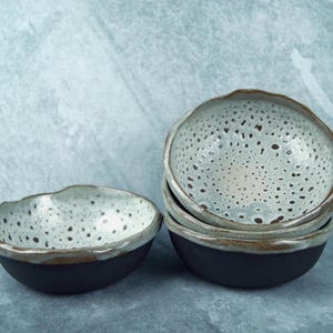 Può includere: Un set di quattro ciotole in ceramica con esterni nero opaco e interni bianchi maculati. Le ciotole presentano bordi organici irregolari con un bordo in argilla marrone visibile, creando un contrasto con la base scura.