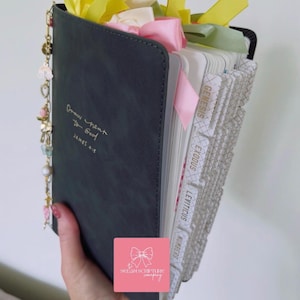 May include: A dark gray Bible with gold lettering that reads "Draw near to God" and "James 4:8". The Bible has colorful ribbons and a charm attached. The pages are labeled with book names.