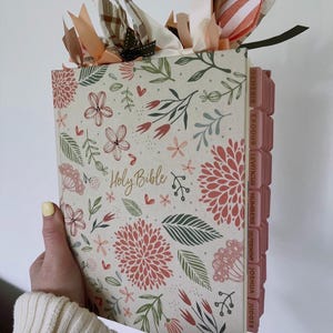 May include: A Holy Bible with a cream-colored cover featuring a floral pattern in shades of pink, green, and brown. The book includes pink index tabs labeled with book names and decorative fabric ribbons tied to the top of the spine.