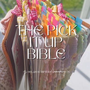 May include: A Bible with a tan cover featuring numerous colorful page tabs and small fabric ribbon bows along the top edge. The text THE PICK IT UP BIBLE and the Selah Scripture Company logo are printed in white on the front of the book.