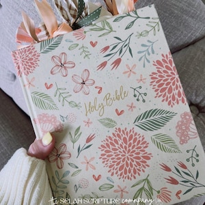 May include: A floral-patterned "Holy Bible" with a cream-colored cover, adorned with pink and peach flowers, green leaves, and the text "Holy Bible". The book has decorative ribbon bookmarks in various colors. The Selah Scripture Company logo is visible.