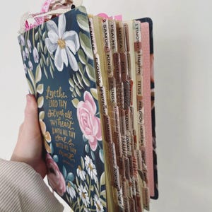 May include: A floral-covered Bible with a navy blue cover, featuring white daisies and pink roses. The cover has the text "Love the Lord Thy God with all thy heart & with all thy soul & with all thy mind." The pages are tabbed with book names.